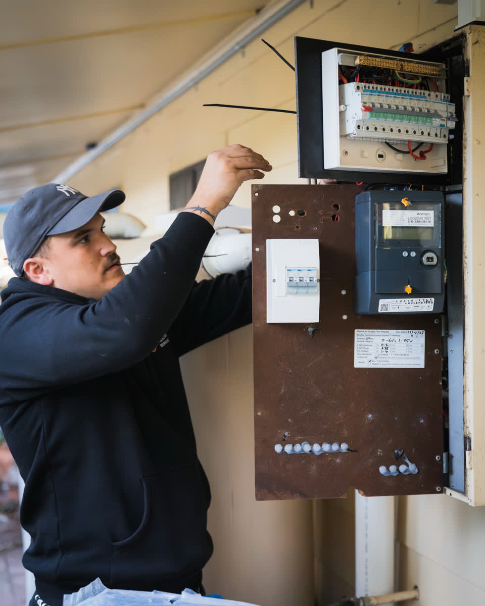 An electrician in a cap working on a meter box and switchboard