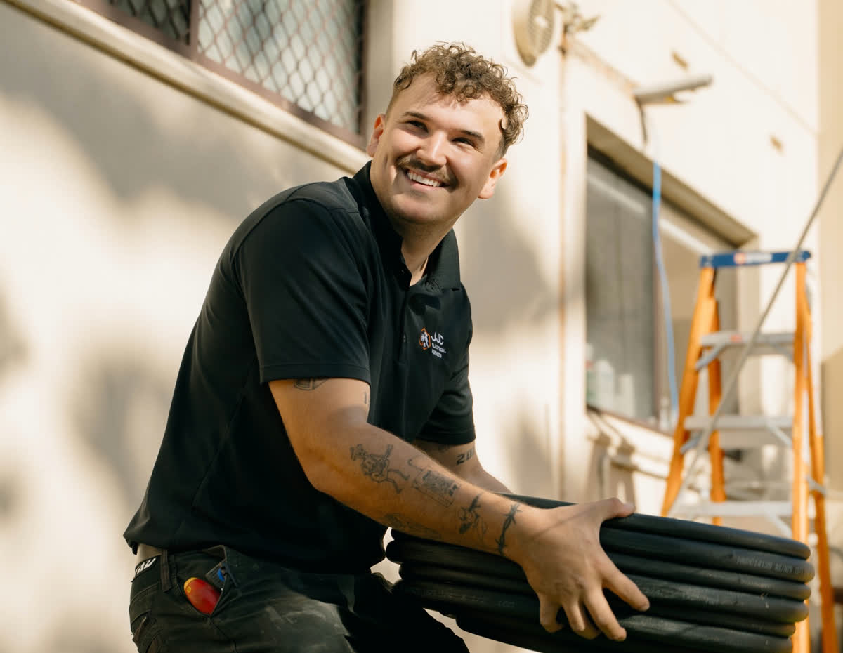 James from JJC Electrical kneeling on a lawn with a coil of conduit, smiling